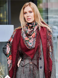 A blond woman wearing an oversized merino wool shawl in rich wine red, decorated with elegant vintage peonies in pink and beige. Designed for wrapping, it combines softness, warmth, and refined floral style.