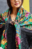 Woman wearing a colorful floral oversized shawl with fringe details against a yellow wall.