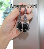 A pair of long black dangle earrings with silver accents, featuring a marcasite design, held in a hand against a white background.