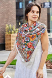 A woman wearing a large white merino scarf adorned with a vintage Slavic herbal pattern. The bright sky blue and yellow details bring a retro folk charm, while the natural sheep fabric and silky fringe add refined softness.