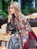 Woman wearing a colorful patterned shawl sitting outdoors. This extra-large wine red shawl features a classic vintage peony pattern in delicate pink and beige tones. Made from natural merino wool, it drapes beautifully as a cozy wrapping scarf.
