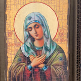 A religious icon painting of the Virgin Mary on a wooden panel, displayed inside a cardboard box. The icon features Mary with a teal-blue head covering and a red dress, set against a gold background with a red halo.