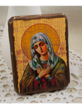 A rectangular religious icon plaque depicting the Virgin Mary with a gold background and brown border, placed on a white lace doily with a soft white backdrop.