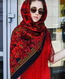 Woman wearing a red floral head shawl with large roses and silky red fringe standing in front of a glass door. Close-up detail of a woman showcasing a luxurious Slavic-style red merino wool palantine. The oversized shawl features detailed botanical and paisley scrollwork over a black and red base, with a highly textured, flowing red fringe hanging across the V-shaped hem.