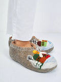 A person wearing brown felted wool slip-on slippers with colorful embroidered winter houses and trees, paired with white pants, on a clean white background.