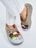A person wearing gray felt house slippers made from natural wool with colorful winter house designs, shown against a plain white background.