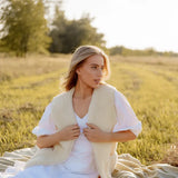 A woman sits on a blanket in a sunlit grassy field, wearing a cream-colored natural sheep wool vest over a white dress. 