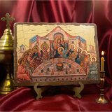 A rectangular wooden Orthodox icon plaque depicting the Last Supper with Christ and apostles, displayed on a small stand against a draped red satin background with a brass candlestick and cross nearby.
