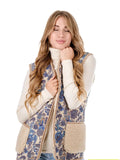 A woman wearing a sleeveless patterned vest with beige sherpa lining over a cream turtleneck and light blue jeans, shown against a plain white background.