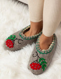 A person wearing gray felt slippers with red rowan berry and green leaf embroidery, standing on a white faux fur rug.