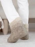 A close-up of a person wearing soft, sheep felt slip-on slippers in a light beige color, paired with white pants, standing on a light-colored floor.