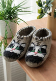 A pair of felted wool slipper shoes shaped like a black-and-white cat face, resting on a wooden table with potted plants in the background.