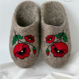 A pair of handmade felt warm slippers in a warm taupe-brown color, decorated with embroidered red poppies and green leaves on the front, set against a white fabric background.
