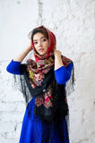 An elegant product lifestyle shot featuring a woman wrapped in a thick, cozy, and completely opaque woolen shawl. The garment showcases a classic Slavic heritage design, splitting into a vibrant red patterned top section worn over her head and a contrasting black bottom section adorned with bold, layered floral rose motifs.