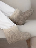 A close-up of a person's feet wearing thick, natural felt boots slippers in a light beige color, resting on a cream-colored armchair.