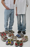 Two people wearing jeans with a collection of embroidered natural woolen slippers in front on a white background.