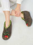 A person wearing brown felt slip-on slippers with green trim and embroidered red-and-yellow fruit motifs, seated on a light neutral floor.