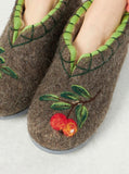 A close-up of a pair of brown wool felt slippers decorated with green leaf embroidery and red/orange berry motifs, shown on feet against a light gray background.