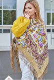 Woman wearing a colorful floral-patterned fringed shawl with a yellow sweater and white pants.