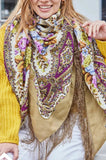 Woman wearing a colorful patterned scarf with a yellow sweater and white pants, holding a product packaging.