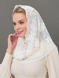 A woman wearing a white lace italian bridal headscarf with floral embroidery and a beige long-sleeve blouse, photographed against a solid gray studio background.