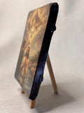 A small rectangular religious art icon  showing a biblical scene, displayed upright on a miniature wooden easel against a plain light background. Theophany Christian artwork showing God the Father and the Holy Spirit dove in a golden glow. Close-up of a rustic wood block icon featuring Jesus in the Jordan River with gold leaf details.