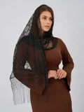 A woman wearing a brown ribbed long-sleeve dress and a sheer black lace italian style head cover with fringe, photographed against a light gray studio background.