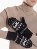 A close-up of a person wearing a beige ribbed turtleneck sweater and holding a pair of black knit mittens with a white Nordic reindeer, a polar bear and pine tree pattern, against a clean white background.