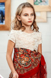 Young girl wearing a white lace top and red patterned Slavic shawl indoors. A close-up view of a high-quality merino wool shawl swatch, showcasing paisley lines and vintage blooming floral patterns over a rich red base.