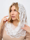 A woman wearing a white lace bridal veil draped over her head and shoulders, paired with a beige top, against a light gray studio background.
