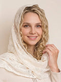 A smiling blonde woman wearing a sheer ivory lace headscarf or shawl draped over her head and shoulders, posed against a soft beige studio background.