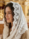 A woman wearing a white lace-trimmed veil over her head and shoulders, paired with a brown robe, photographed indoors in a softly blurred church-like setting. 