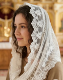 A woman wearing a white lace-trimmed veil over her head and shoulders, paired with a brown robe, photographed indoors in a softly blurred church-like setting. A circular badge in the lower right reads 'Мнедового питания'.