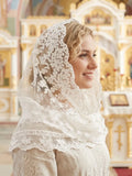 A woman wearing a white lace veil over her head and shoulders, standing in a brightly lit church interior with ornate gold and cream architectural details in the background.