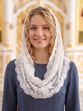 A smiling blonde woman wearing a white lace-trimmed head covering over a navy blue dress, set against a softly blurred ornate interior background.
