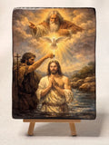 A rectangular wooden icon of a religious painting depicting Jesus in water with a dove and God above, displayed on a small wooden easel against a light gray background. Catholic devotional painting of the Baptism of Christ on a solid wood block panel. Traditional style Catholic wood art block with rustic dark edges for a home prayer altar.