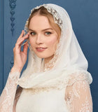 A woman wearing a white lace bridal veil with floral embroidery and a matching long-sleeve lace dress, posed against a blue paneled background. 
