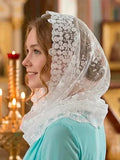 A woman in profile wearing a white lace-trimmed sheer floral head covering over her hair, with a turquoise top, photographed indoors with warm blurred candlelight in the background.