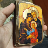 A hand holding a small wooden religious Orthodox icon featuring a painted Jesus family scene with gold halos and dark robes, photographed indoors against a blurred background.
