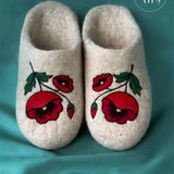 A top-down view of premium handcrafted women's house slippers made from thick, un-dyed cream white sheep wool felt. Both slippers are beautifully adorned with an artisan needle-felted design of a large blooming red poppy flower accompanied by smaller buds, green leaves, and delicate black stems.