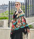 A woman with a large winter shawl of natural merino wool elegantly styled over her shoulders and covering her body till knees displaying traditional vintage patterns of pink peonies, bright wildflowers, and a black border with a long fringe.