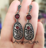 A pair of long sterling silver Armenian earrings with floral ethnic patterns and lab garnet stones, held in a hand.