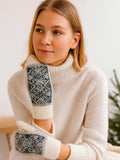 Fluffy white wool mittens featuring an intricate grey and black Nordic geometric snowflake pattern across the back of the hand. A model wears them crossed over a glossy metallic green down winter jacket.