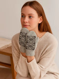 A woman wearing a cream knit sweater holds up a pair of light gray knit  woolen mittens with a dark gray geometric snowflake  Nordic pattern on the palms.