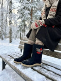  A close-up lifestyle shot of a woman sitting on a snow-dusted wooden park bench in a winter forest, wearing fashionable black Slavic felt boots. The boots feature a modern beige and gray rubber outsole with a thick tread, protecting the handcrafted black sheep wool upper from the cold ground.
