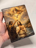 A hand holding a small rectangular leather-bound icon with a religious painting on the cover, showing an angel, a dove, and two figures in water. Handmade religious gift showing John the Baptist pouring water on Jesus on a wooden display easel. Premium double-lacquered wood panel art piece of the Holy Trinity baptism scene on a tabletop. Gilded religious icon of the Epiphany of Christ protected by durable clear lacquer coats.