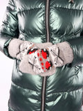 A close-up of a person wearing gray knit winter gloves with a red and black pattern, holding them in front of a shiny dark green puffer jacket. Thick knit mittens with an elastic ribbed cuff designed to accommodate flexible hand circumferences ranging accurately from 6 to 8 inches. The traditional European folklore motif includes detailed red berries, black-and-white accents, and geometric shapes.