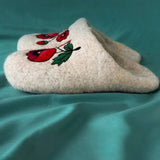 Close-up of extra comfortable felted wool slippers displaying a thick, seamless construction without external outsoles. The pristine white felt surfaces serve as a striking canvas for a vibrant red and black poppy floral arrangement hand-stitched near the foot openings.
