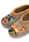 Handcrafted sheep wool slippers in a cozy gray melange shade, designed with a festive autumn and winter theme. The low-cut slipper shoes feature unique hand-guided felt applications, with a cheerful orange squirrel motif on the right foot and a bunch of bright red berries on a branch on the left foot, accented by an orange stitched trim along the collar.