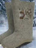 A pair of tall beige felt winter boots with brown floral embroidery on the side, standing in snow with a blurred snowy outdoor background. Close-up of a stylish winter outfit featuring traditional Slavic-style wool felt boots reimagined with a modern twist, set against a background of frosty, snow-laden trees. 